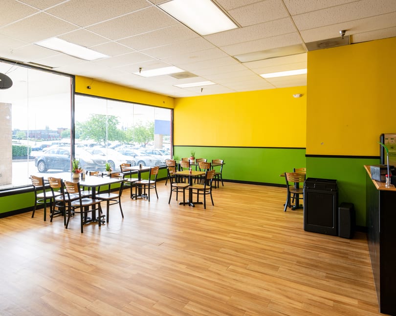 Empty restaurant dining room with bright yellow and green walls, wooden floors, and tables by large windows.