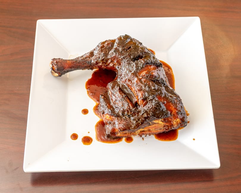 A large piece of dark, glazed jerk chicken with sauce on a white square plate.