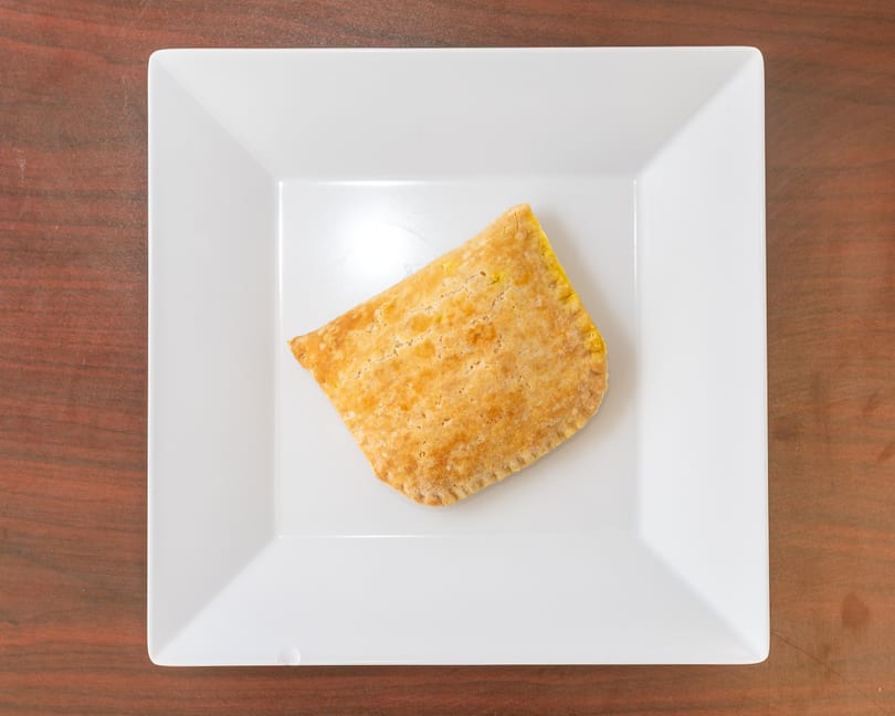 Golden brown Jamaican beef patty on a white square plate, centered on a wooden table.