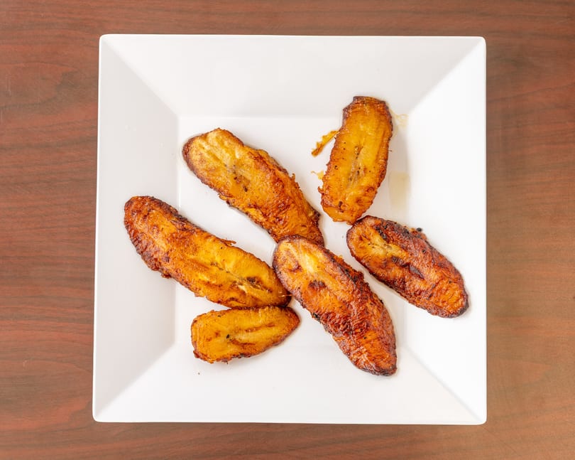 Six golden-brown fried plantains are arranged on a white square plate, on a wooden table.