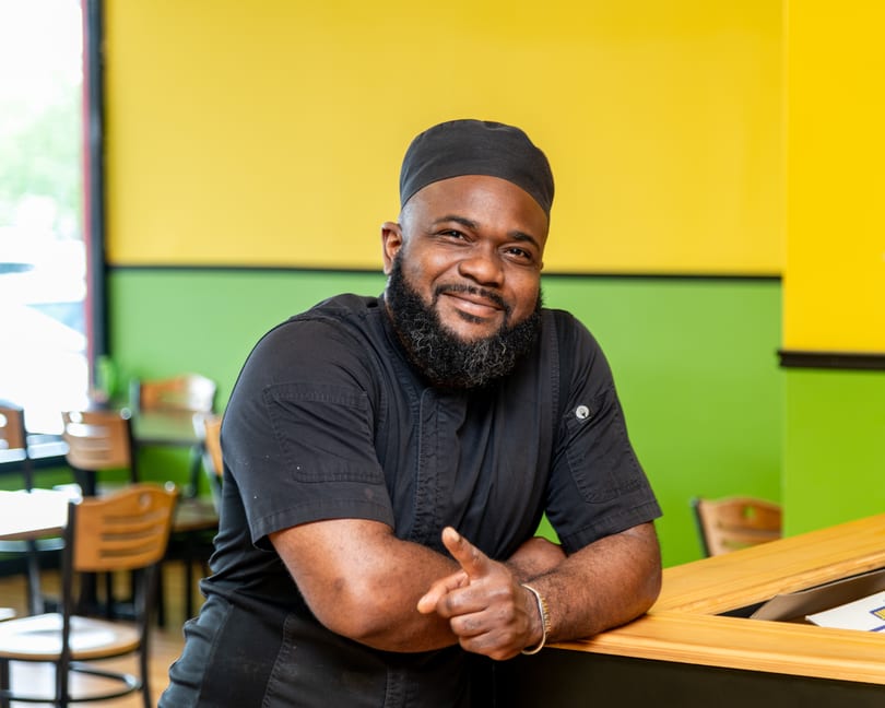 Smiling chef in black uniform and cap leaning on a wooden counter, pointing forward.