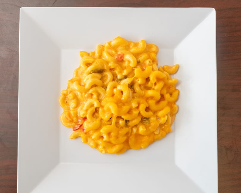 Creamy macaroni and cheese with red and green peppers on a white square plate.